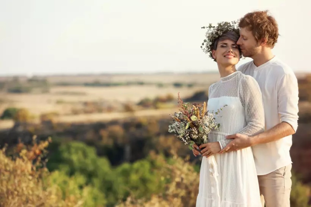 Jeune couple de mariés dans un champ, la mariée en robe fluide et couronne de fleurs, tenant un bouquet champêtre, le marié l’embrassant tendrement, symbole d’un mariage champêtre chic et naturel.